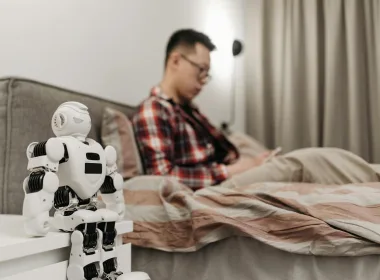 A modern bedroom featuring a robot on a bedside table and a man sitting on a bed.