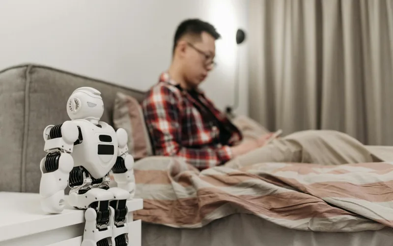 A modern bedroom featuring a robot on a bedside table and a man sitting on a bed.